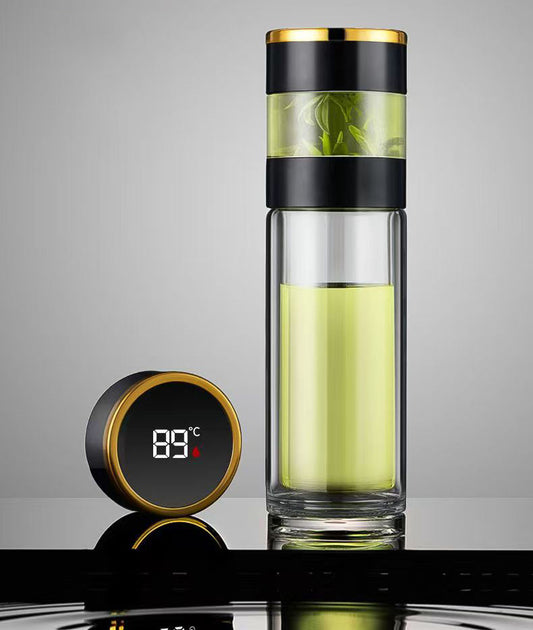 The Smart water boatel, featuring a black and gold lid and filled with light green liquid, stands beside a digital thermometer showing 89°C on a reflective surface against a gray background—ideal for hydration tracking.